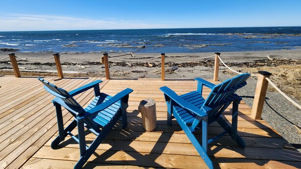 Een houten terras met twee blauwe Adirondack-stoelen, gericht op een rotsachtig strand en de oceaan.