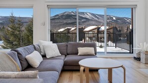 Ett modernt vardagsrum med en grå hörnsoffa, ett runt soffbord och stora fönster med utsikt över ett snötäckt berg och byggnader.
