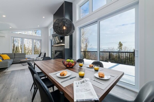 Un comedor moderno con una mesa de madera, sillas y un periódico. Hay un balcón con barandilla y vistas a árboles.