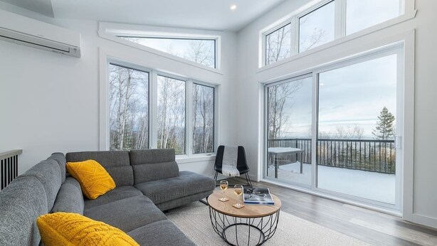 A modern living room with a grey sectional sofa, a round coffee table, and large windows offering a view of trees and a balcony. - Hôtel à la maison - Le Quintessence, spa et vue (Saint-Irénée)