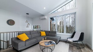 A modern living room with a grey sectional sofa, a round coffee table, and a black chair. There are large windows with a view of trees, a clock on the wall, and pendant lights. - Hôtel à la maison - Le Quintessence, spa et vue (Saint-Irénée)