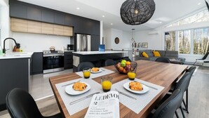 A modern kitchen with dark cabinets, a wooden dining table with breakfast plates, and a hanging light fixture. - Hôtel à la maison - Le Quintessence, spa et vue (Saint-Irénée)