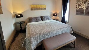 Ein Schlafzimmer mit einem Bett, zwei Nachttischen, einer Bank, einem Fenster mit Vorhängen und Wandbildern.