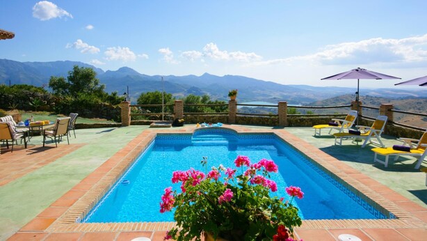 Une piscine rectangulaire à l’eau d’un bleu limpide, bordée d’une margelle en pierre. Des chaises longues et des parasols sont disposés autour de la piscine, et on aperçoit des montagnes au loin.