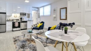 Une cuisine moderne avec des meubles blancs, un four à micro-ondes et une cuisinière. On y trouve un coin repas avec une table ronde et des chaises, ainsi qu’un canapé avec des coussins.