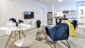 Une cuisine moderne avec des meubles blancs, un four à micro-ondes et une cuisinière. On y trouve un coin repas avec une table et des chaises blanches, ainsi qu’un téléviseur fixé au mur.