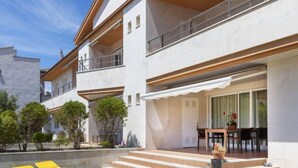 A modern two-story building with a covered outdoor seating area, steps leading to a patio, and a small garden with yellow lounge chairs. - Gabriel by Interhome (Sant Jordi)