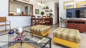 A modern living room with a glass coffee table, yellow ottomans, a dining area with wooden chairs, a large flat-screen TV, and a kitchen visible through an open door. - Gabriel by Interhome (Sant Jordi)