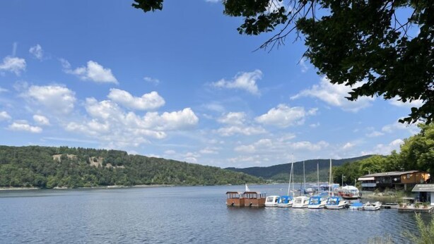 Un lac paisible où sont amarrés des bateaux le long de la rive, entouré de collines et d’arbres.