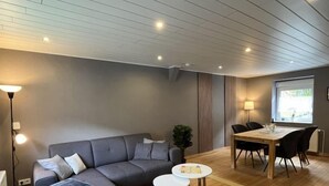 Un salon moderne avec un canapé d’angle gris, une table à manger avec des chaises et une petite table ronde sur laquelle est posé un jeu de société.