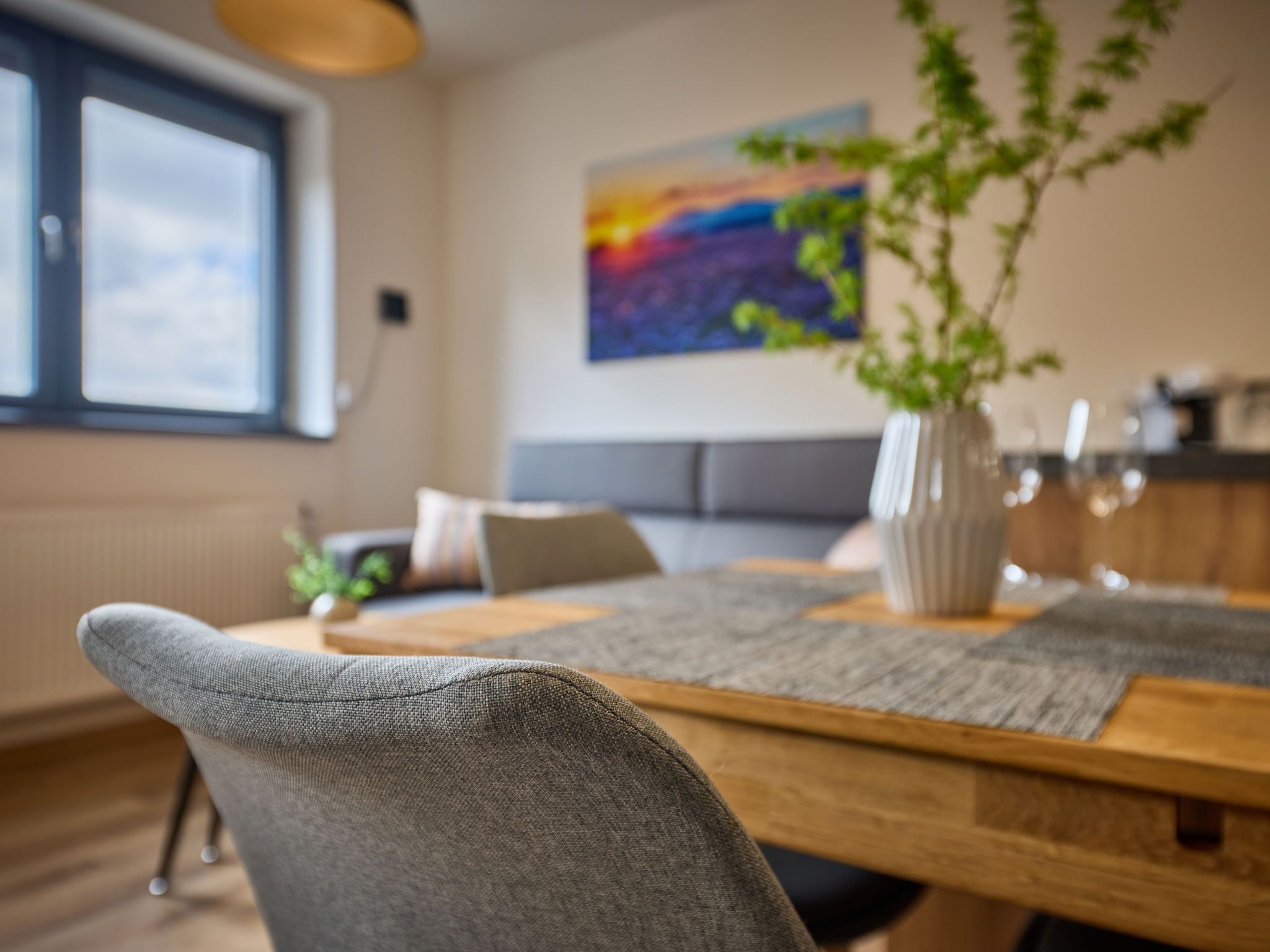 A modern dining area with a wooden table, grey upholstered chairs, a vase with a plant, and a colorful abstract painting on the wall.