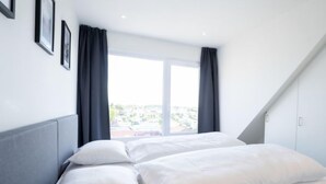 Quarto moderno com uma janela grande, cama com roupa de cama branca e cabeceira cinzenta.
