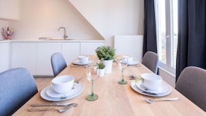 Uma sala de jantar com uma mesa posta para quatro pessoas, uma cozinha ao fundo e vista para o exterior através de grandes janelas.