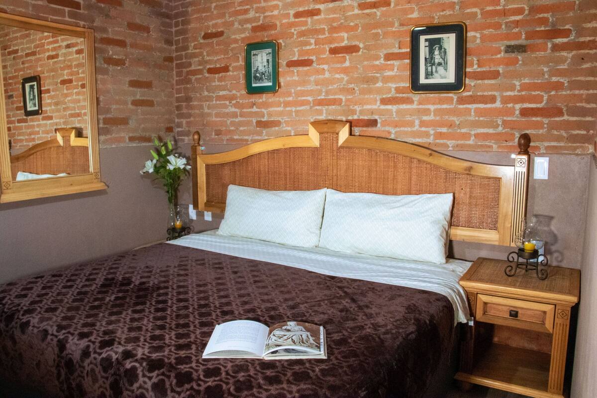 Un dormitorio con pared de ladrillo, cabecera de madera, cama con colcha estampada, mesita de noche con lámpara y espejo en la pared.