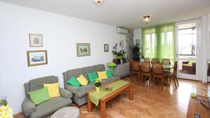 Ein Wohnzimmer mit einer Sofagarnitur, einem Esstisch mit Stühlen und einem Couchtisch. An der Wand befinden sich gerahmte Bilder, eine Topfpflanze und ein Fenster mit Vorhängen.