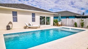 Una piscina rectangular con superficie de agua azul cristalina, rodeada por una zona de patio de tonos claros con una tumbona y una mesita.