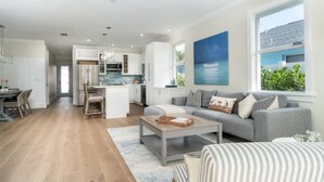 Una sala moderna con un sofá gris, una mesa de centro y una silla a rayas. Se ve una cocina al fondo con armarios blancos y vistas al exterior.