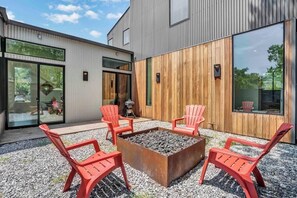 A modern outdoor seating area with four red chairs around a square fire pit, set against a building with large windows and a wooden wall.