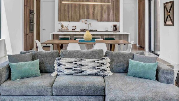 Uma sala de estar moderna com um sofá seccional, uma mesa de centro e uma cozinha ao fundo.