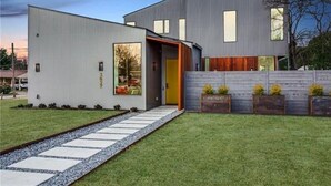 Um edifício moderno de dois andares com uma grande porta de vidro, um caminho pavimentado que leva à porta e uma pequena área de jardim com plantas em vasos.