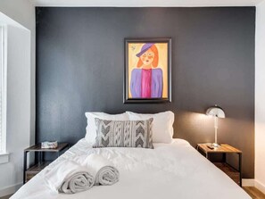 A neatly made bed with white linens, a pillow with a geometric pattern, a framed painting on the wall, and a bedside table with a lamp.