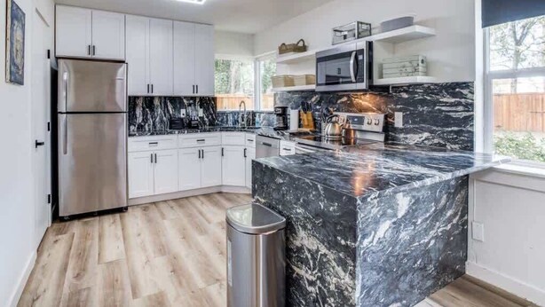 Cozinha moderna com armários brancos, ilha de mármore preto, eletrodomésticos de aço inoxidável e uma janela que traz luz natural.