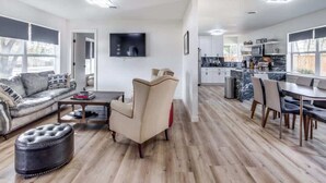 Uma sala de estar moderna com sofá, apoio de pés e mesa de centro, uma área de jantar com cadeiras e uma cozinha com armários e uma televisão.