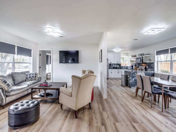 A modern living room with a sofa, ottoman, and coffee table, a dining area with chairs, and a kitchen with cabinets and a TV.