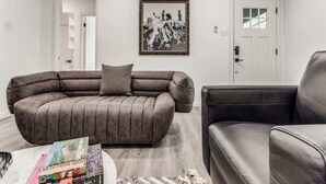 Una sala de estar con un sofá grande en forma de L, un sillón más pequeño, una mesa de centro con un libro y un cuadro en la pared.