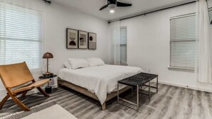 Un dormitorio con cama, una silla, una mesa, ventilador de techo y ventana con persianas.