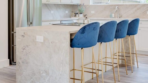Cocina moderna con refrigerador de acero inoxidable, encimeras de mármol, una isla central con taburetes de bar, iluminación colgante y un ventanal amplio.