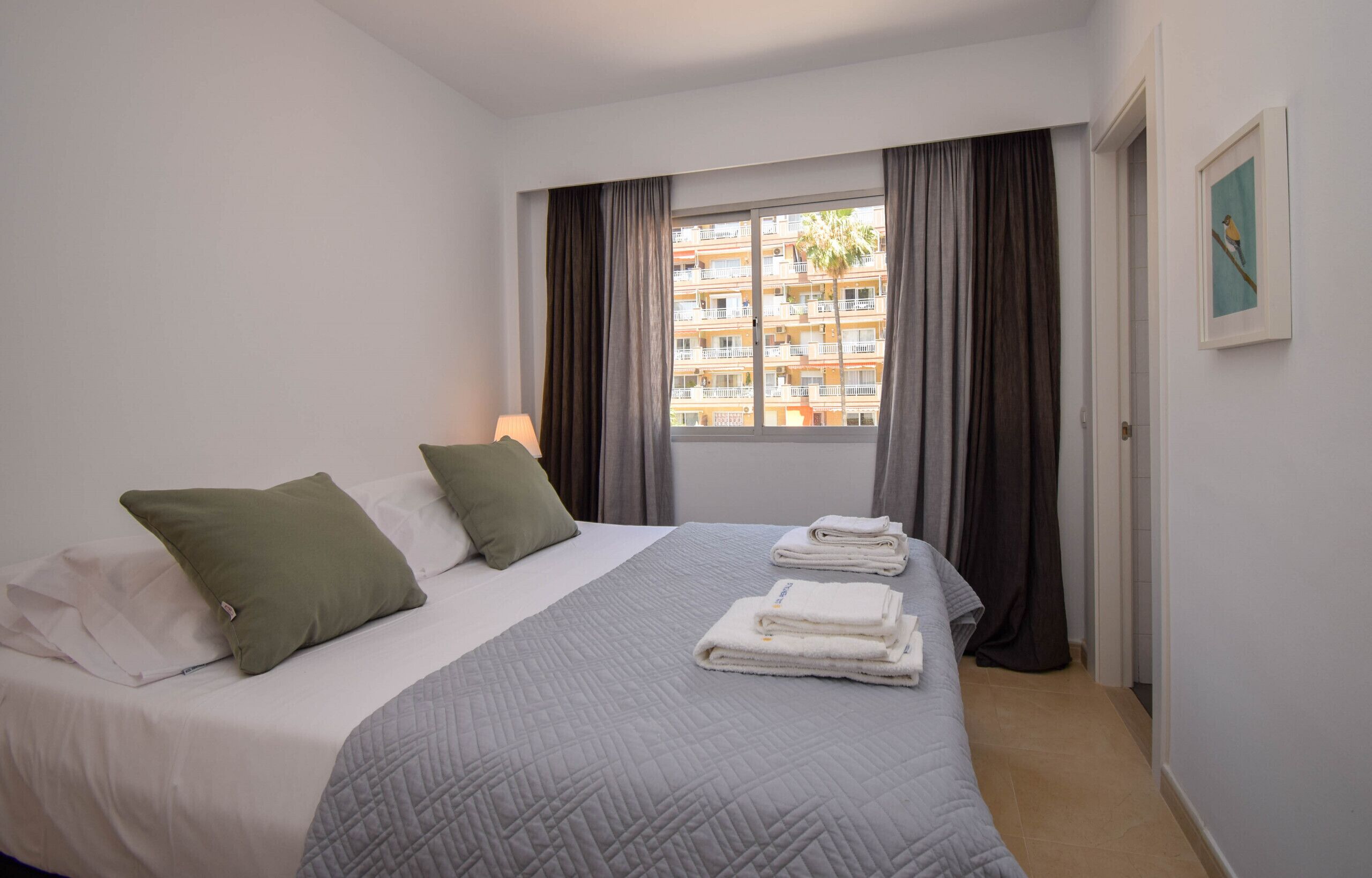 Quarto de hotel com cama, travesseiros, colcha acolchoada, janela com cortinas, quadro na parede e vista para um edifício.
