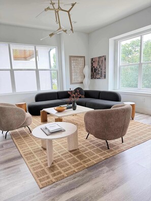 A modern living room with a sectional sofa, two armchairs, a coffee table, and a rug. There are large windows, a ceiling light fixture, and artwork on the walls. - Driem House |Chic | Highland Park Adjacent (Dallas)