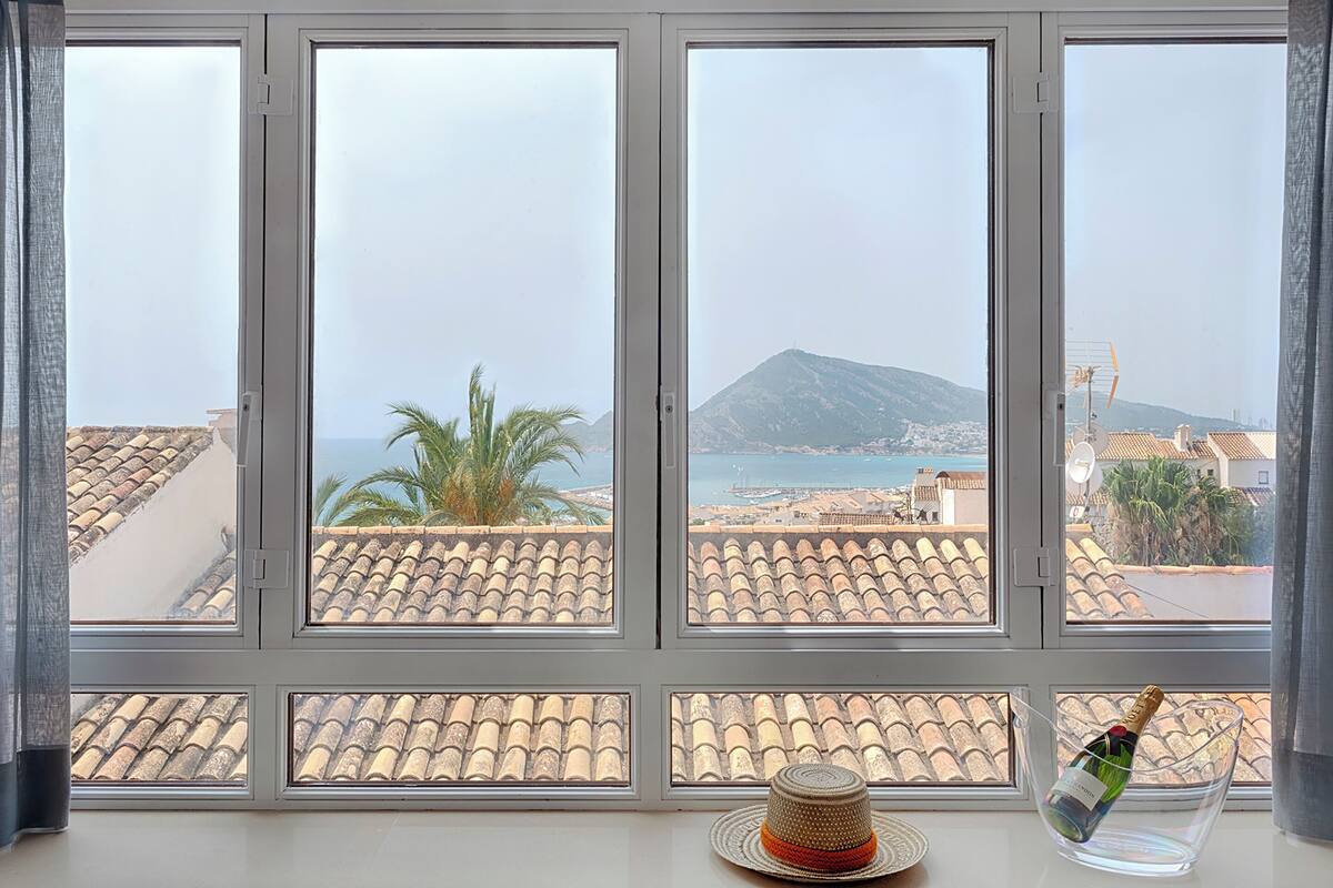 Una ventana con vista a un pueblo costero, una montaña y un sombrero y una botella en el alféizar.