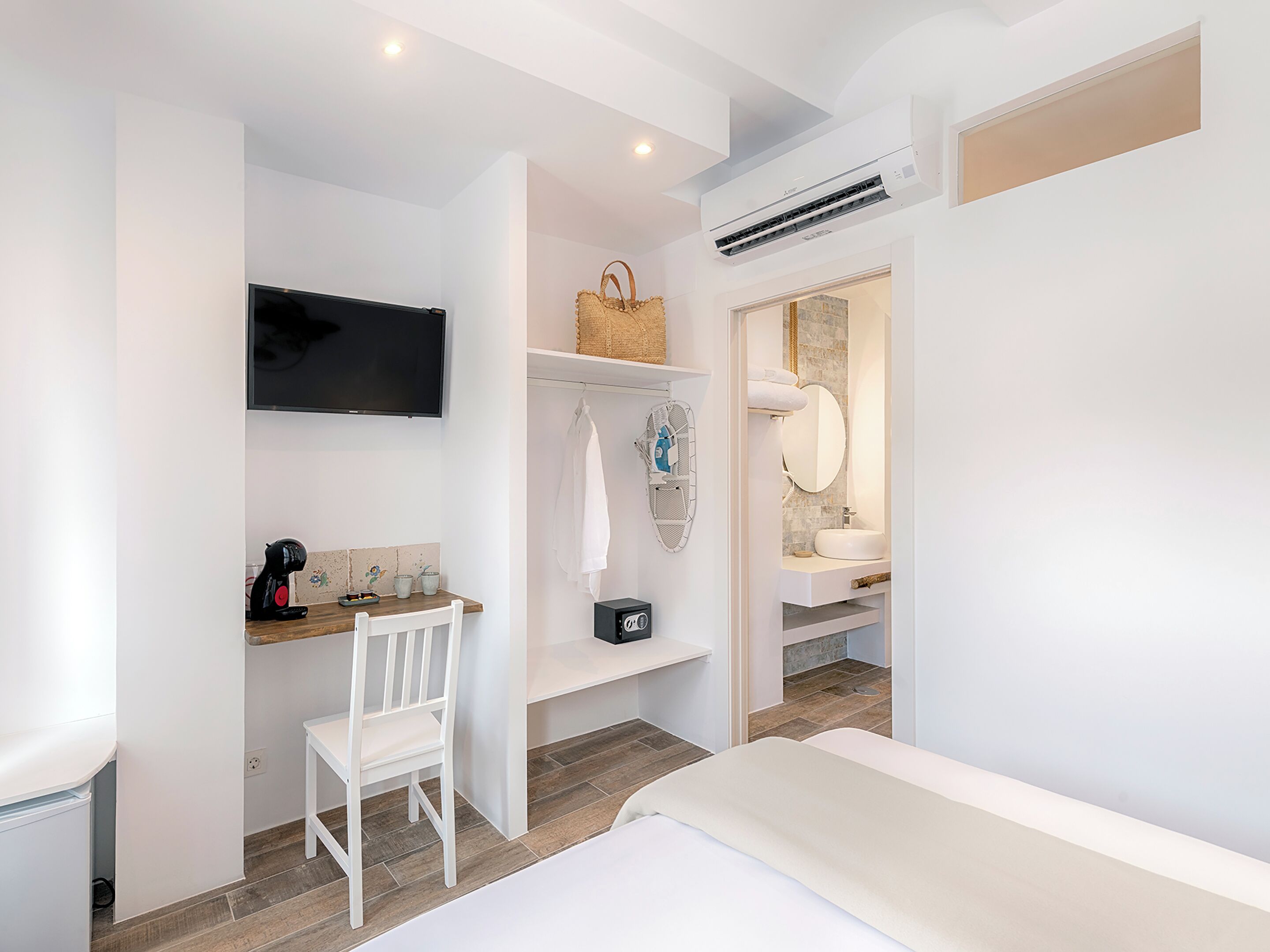 Une chambre d’hôtel comprenant un lit, un bureau avec une chaise, une télévision, une étagère avec un panier, et une salle de bain avec un lavabo et un miroir.