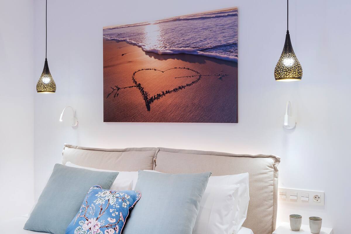 Un dormitorio con cama, almohadas, una mesita de noche con dos tazas y un cuadro en la pared de una playa con un corazón dibujado en la arena.