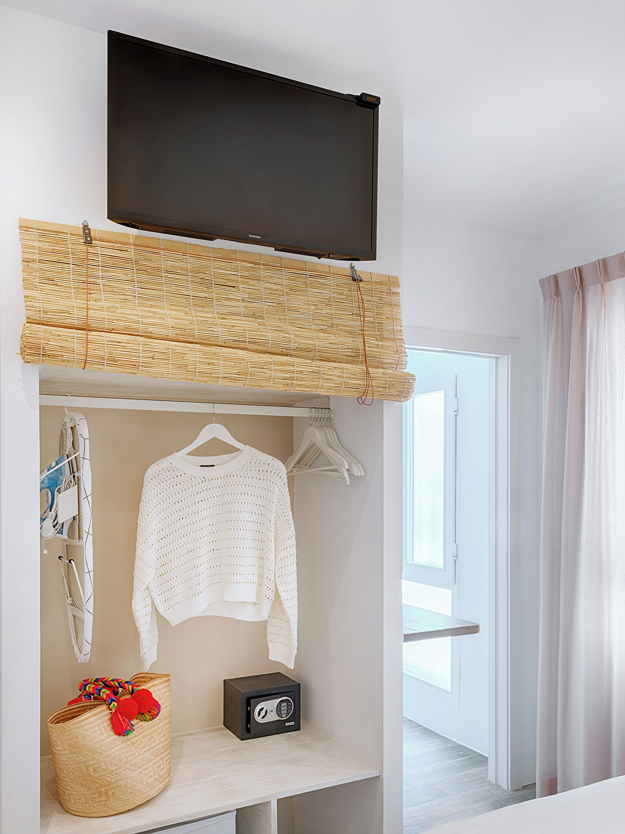 A modern room with a TV mounted on the wall, a woven bamboo blind, a white knitted sweater, a small radio, and a woven basket with colorful items.