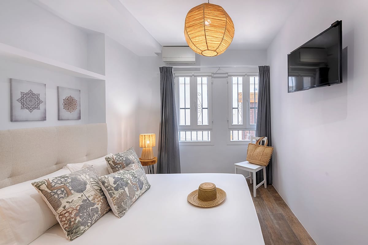Un dormitorio con cama, lámpara colgante, televisor montado en la pared y ventana con cortinas.