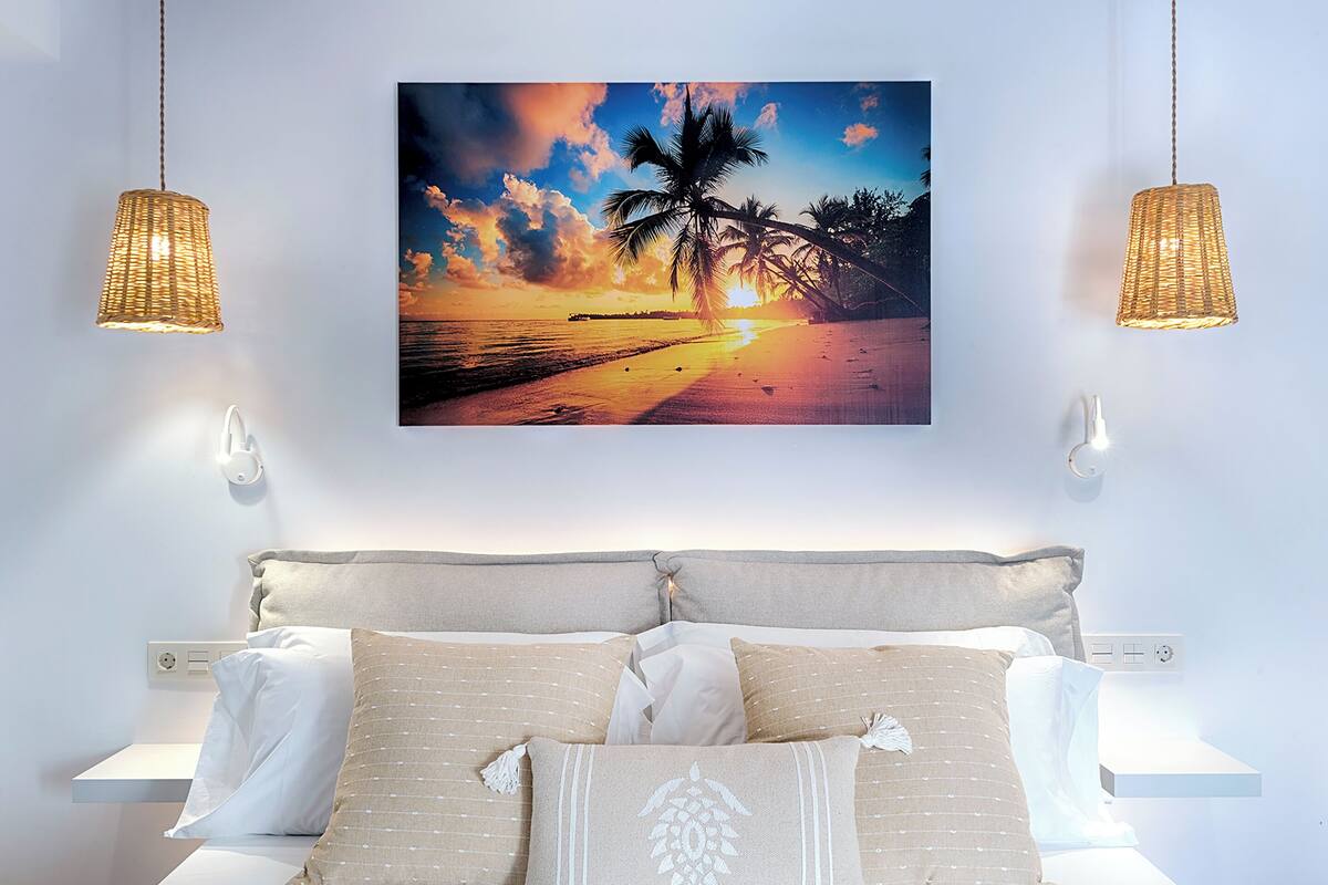 Un dormitorio con una cama, almohadas, un cuadro colgado en la pared de un atardecer y dos lámparas colgantes.