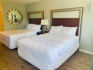 A hotel room with two single beds, a round mirror, a nightstand with a lamp, and a wooden floor. - Ocean One Inn (Sneads Ferry)
