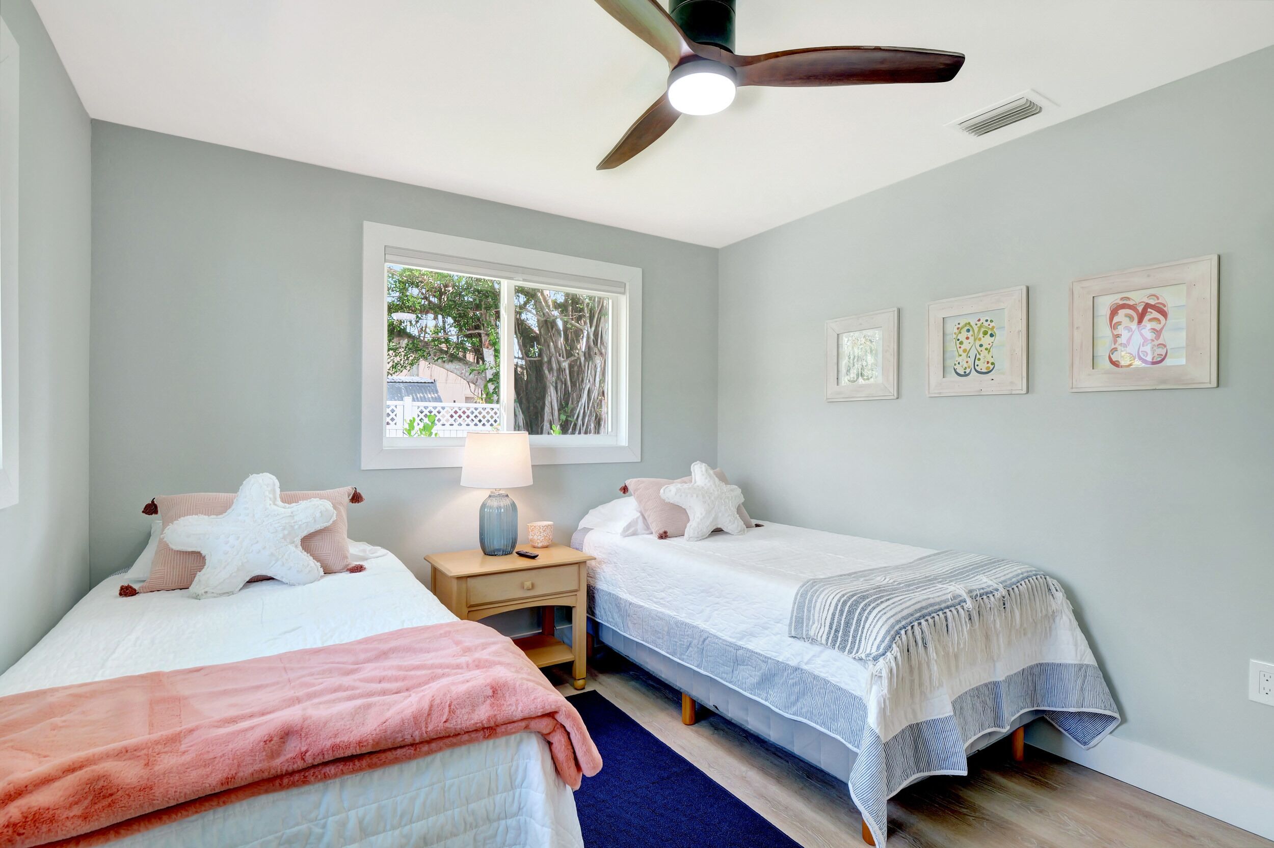 A bedroom with two beds, a ceiling fan, a window, and framed artwork on the wall.