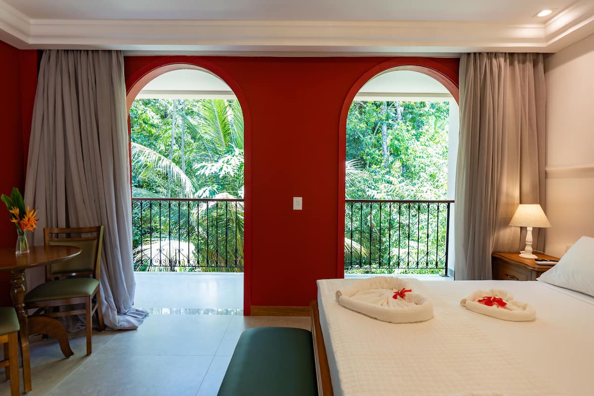 A hotel room with a bed, a desk, and a view of a lush garden through a balcony.