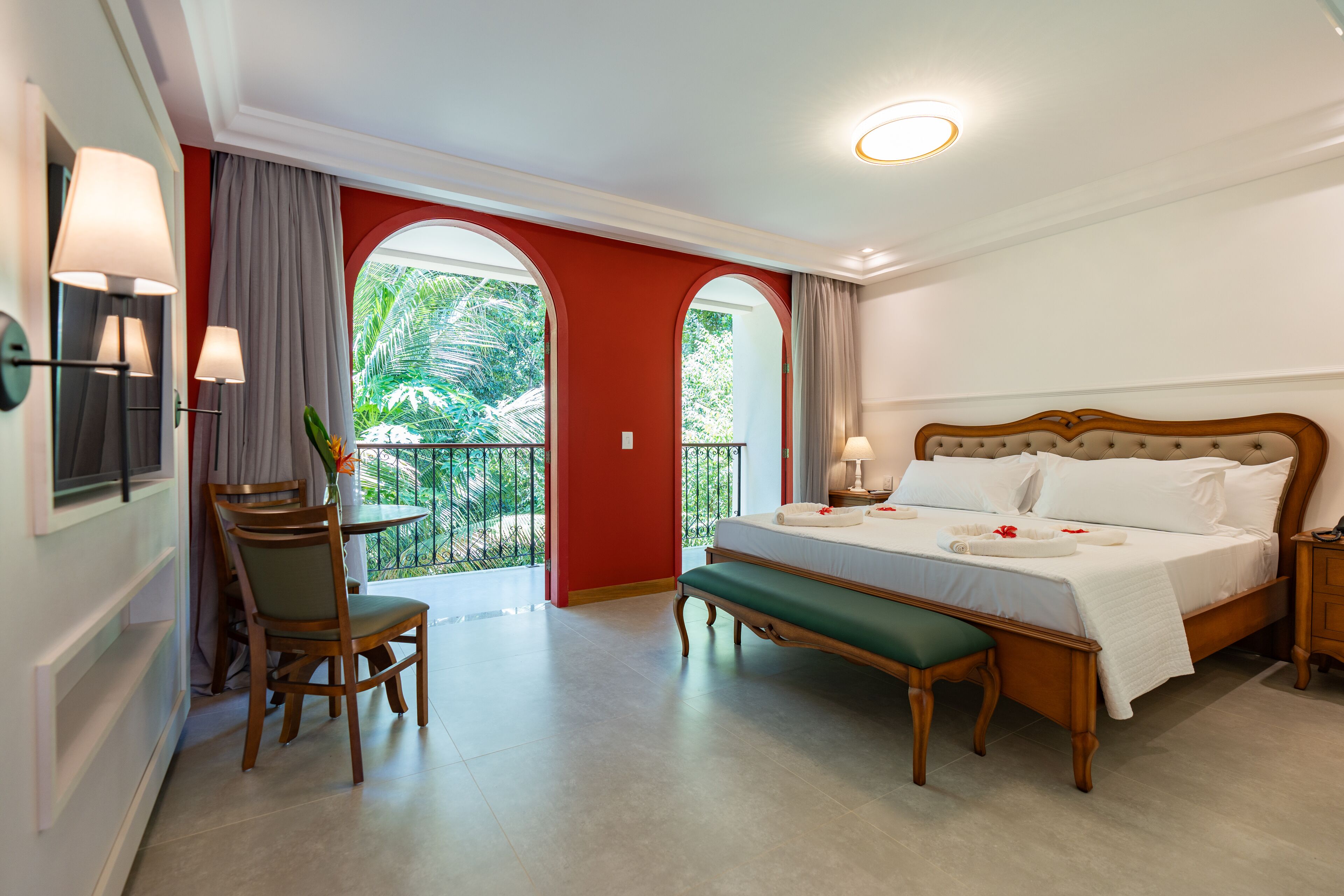 A hotel room with a large bed, a desk with a chair, a small table, and a balcony with a view of greenery.