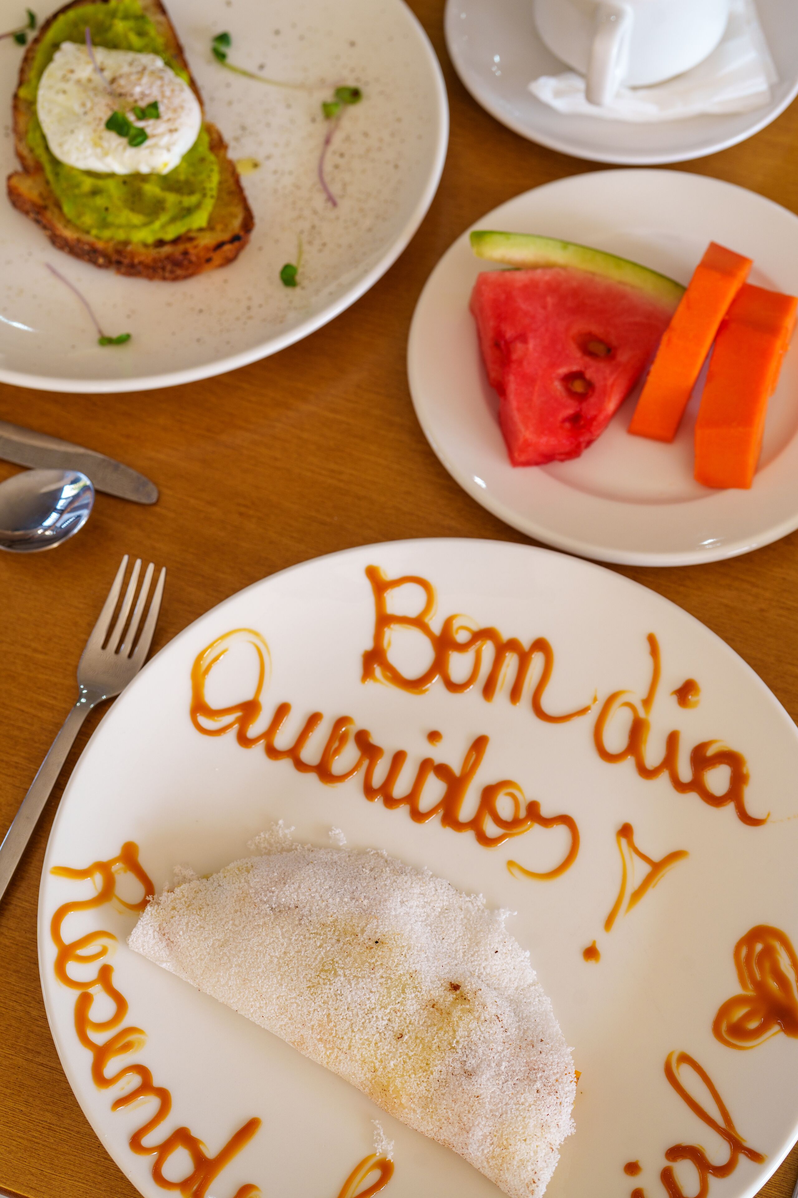 A breakfast plate with a crepe, toast with avocado and egg, and a plate with watermelon and carrot slices.