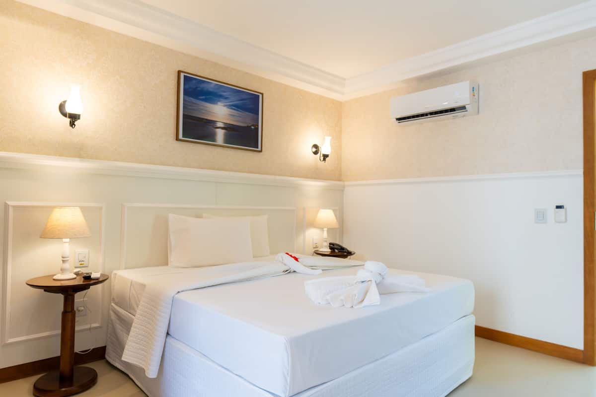 A hotel room with a large bed, two bedside lamps, a painting on the wall, an air conditioning unit, and a telephone on the nightstand.