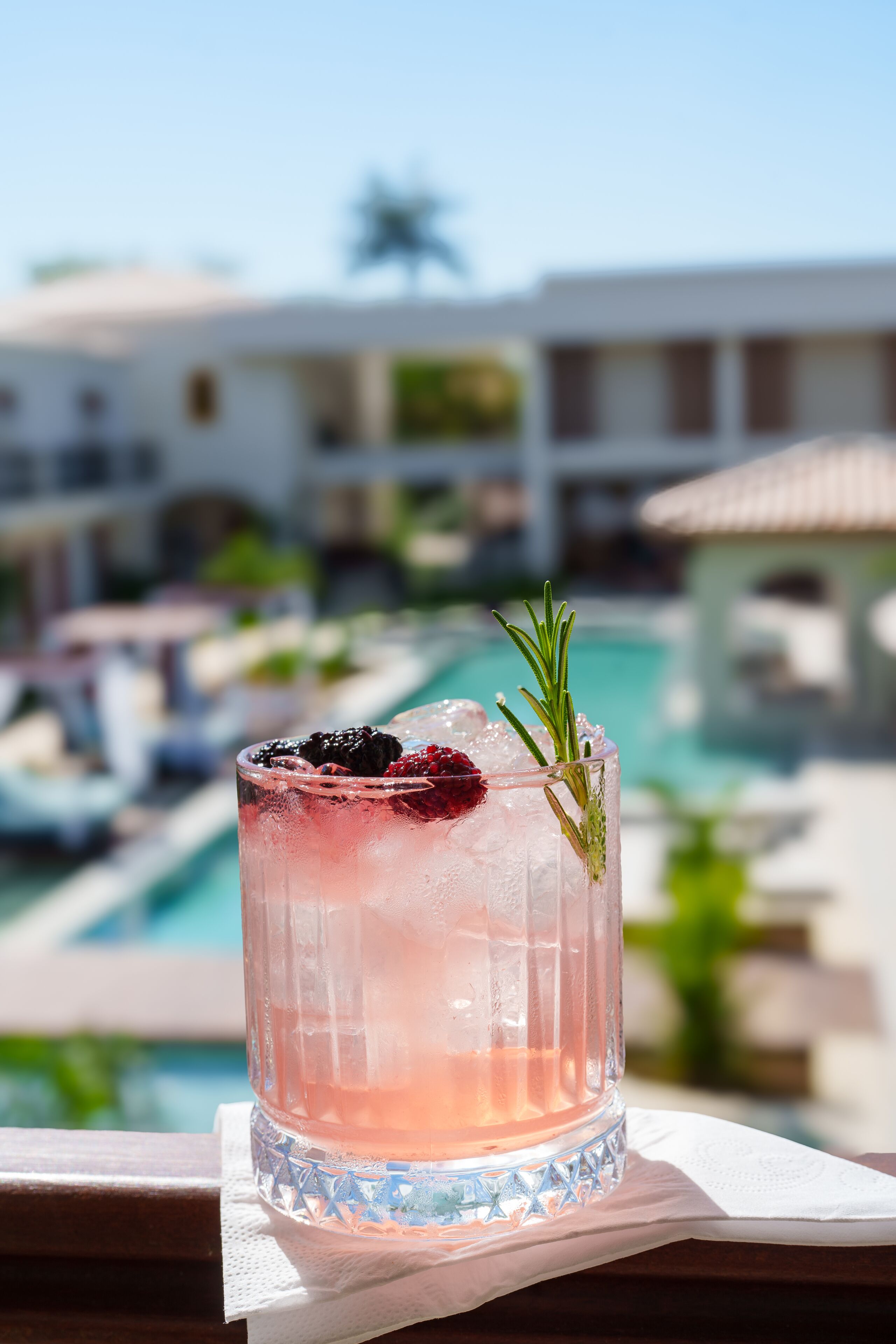 Ein Cocktail mit Garnitur, serviert auf einer Serviette in Poolnähe, mit einem Gebäude im Hintergrund.