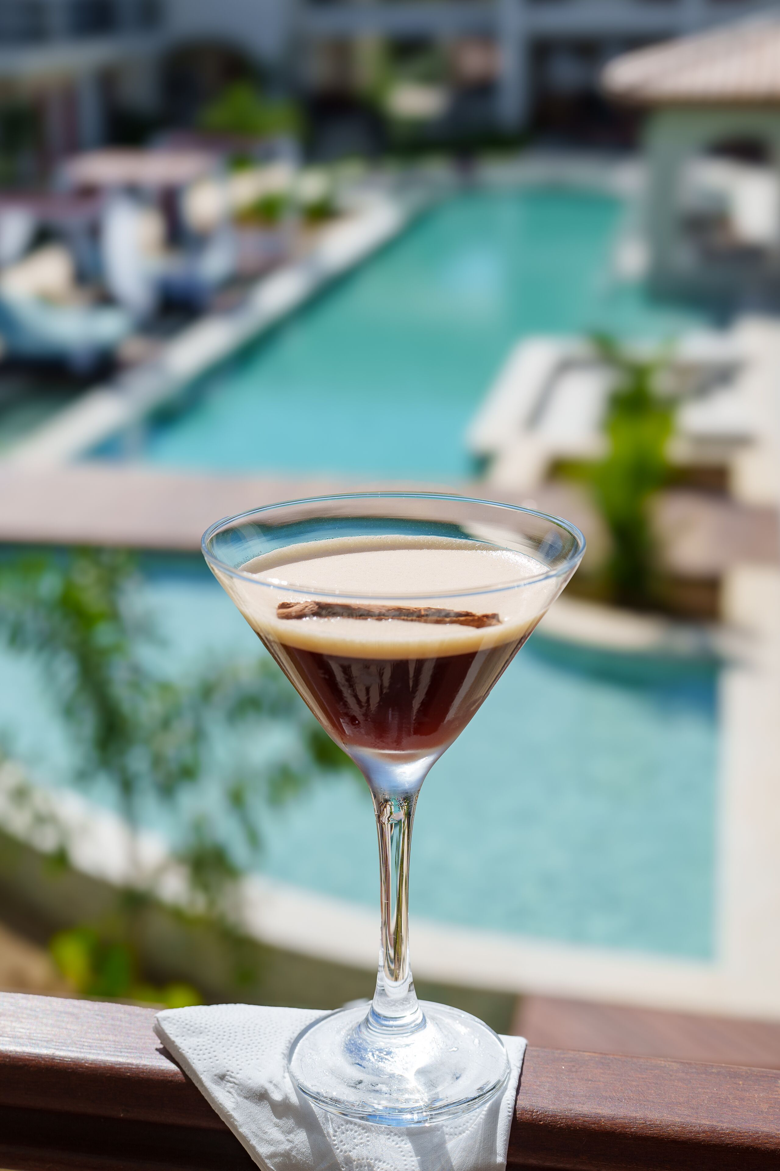 A martini glass with a frothy top on a wooden railing, overlooking a swimming pool with lounge chairs and umbrellas.