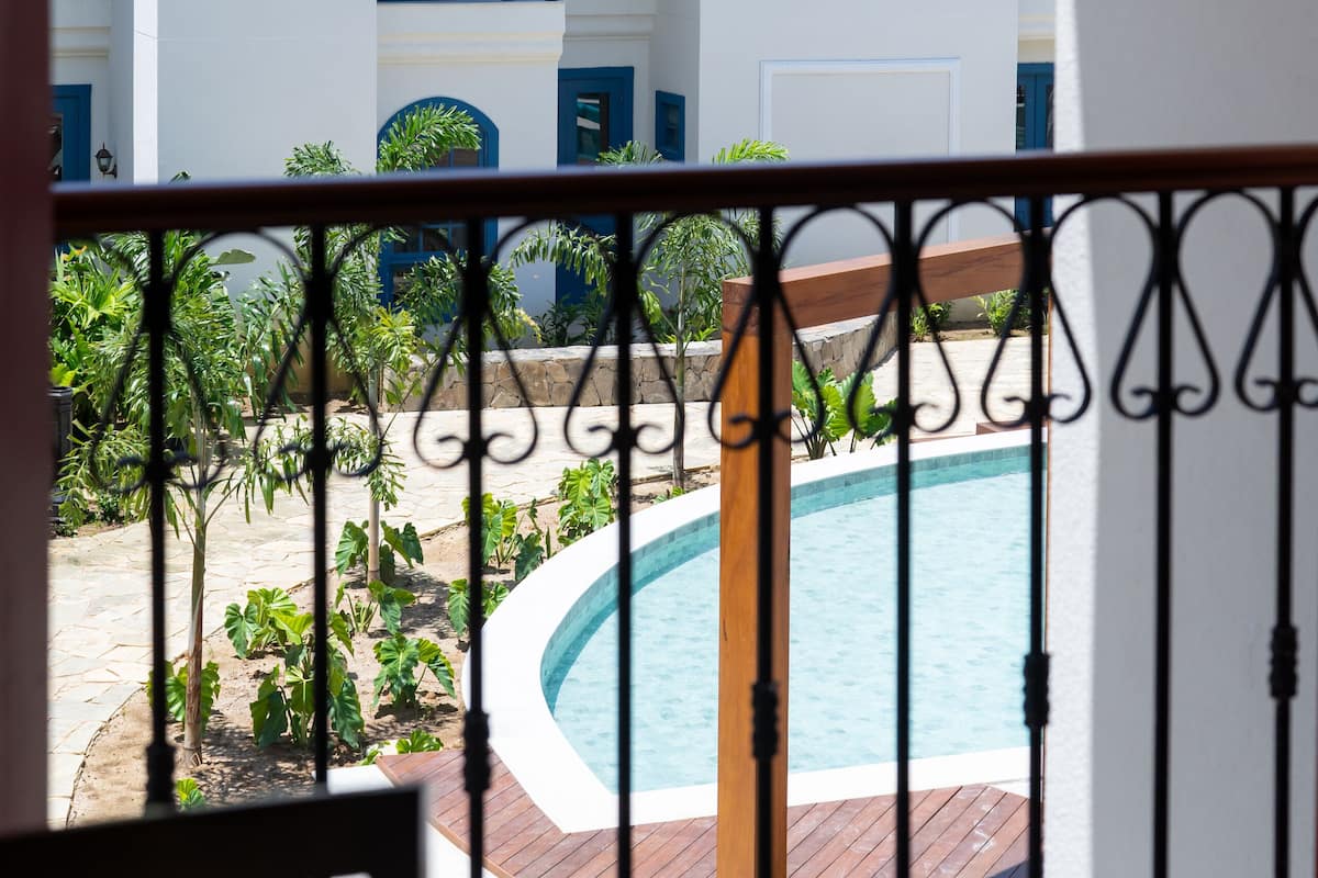 A view of a pool area with a white building in the background, featuring balconies with blue railings and a tiled roof.