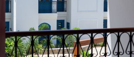 A view of a pool area with a white building in the background, featuring balconies with blue railings and a tiled roof.