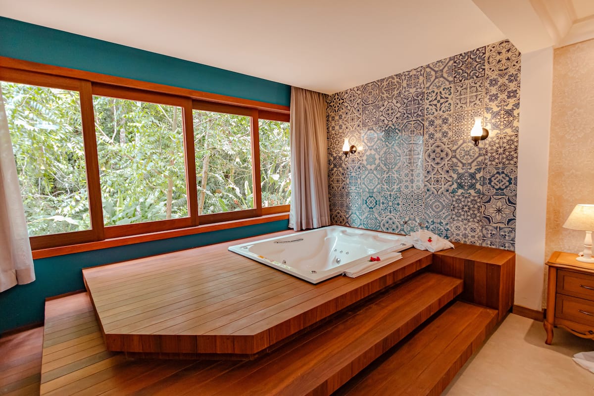 A modern bathroom with a wooden bathtub, a large window with a view of greenery, patterned tiles, and a small bedside table with a lamp.